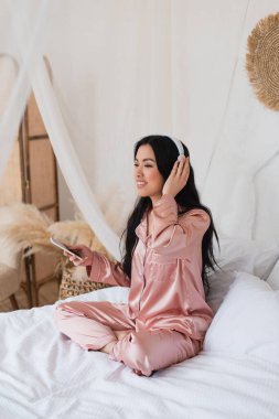 young asian woman in pink silk pajamas sitting on bed with cellphone and touching headphones with hand in bedroom clipart