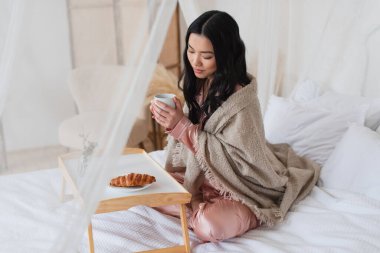 young asian woman in silk pajamas and blanket sitting on bed with crossed legs and having breakfast in bedroom clipart
