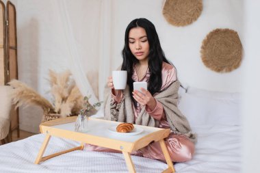 İpek pijama ve battaniyeli genç Asyalı kadın bacak bacak üstüne atmış yatakta oturuyor, kahve içiyor ve yatak odasında cep telefonu tutuyor.