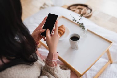 Yatak odasında kahve ve kruvasanla cep telefonuyla mesajlaşan esmer bir kadının kısmi görüntüsü.