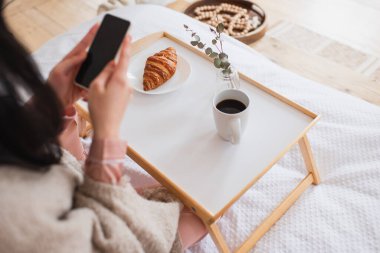 Yatak odasında kahve ve kruvasanla cep telefonu tutan esmer kadının kısmi görüntüsü.