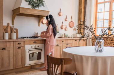 young asian woman in pink silk pajamas and headphones cooking breakfast near table in kitchen clipart