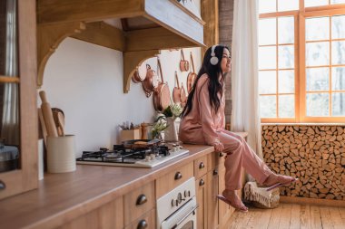 Pembe ipek pijamalı ve kulaklıklı gülümseyen genç Asyalı kadın fırının yanında oturuyor ve mutfaktaki müziğin tadını çıkarıyor.