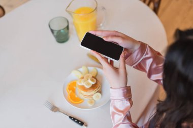 Genç bir kadının elinde akıllı telefon tutarken ve mutfakta portakal suyunun yanında meyveli krep çekerken kısmi görüntüsü.