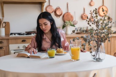 İpek pijamalı genç Asyalı kız mutfakta kahvaltı ederken kitap okuyor.