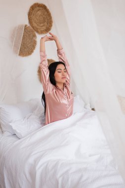 Uykulu, ipek pijamalı genç Asyalı kadın uzanmış elleriyle ve yatak odasında kapalı gözlerle uyanıyor.