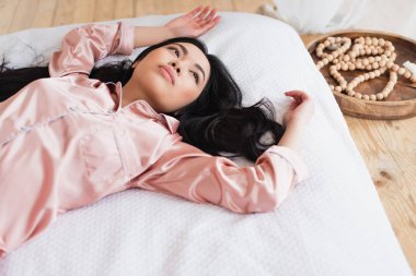Pijamalı, beyaz çarşafların üzerinde uzanmış, yatak odasında uzanmış Asyalı bir kadının yüksek açılı görüntüsü.