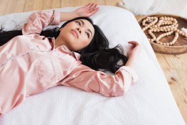 high angle view of young asian woman in pajamas lying on white linen with outstretched hands in bedroom clipart