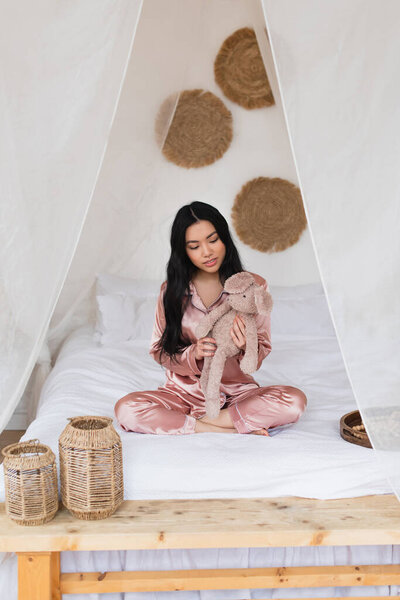 young asian woman in silk pajamas sitting with crossed legs and holding soft toy in bedroom