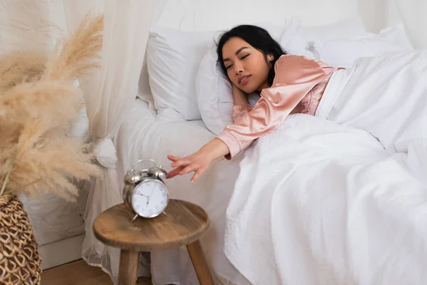 年轻的亚洲女人躺在白色的亚麻布上 手伸向床头柜 桌上摆放着闹钟 — 图库照片