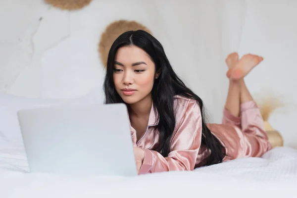 young asian woman in silk pajamas lying on bed and looking at laptop in modern bedroom