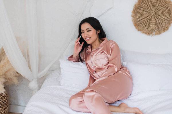 happy young asian woman in pink silk pajamas lying on bed and speaking on cellphone in bedroom