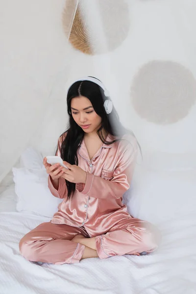 Молода Азіатка Рожевій Шовковій Піжамі Сидить Ліжку Навушниками Мобільним Телефоном — стокове фото
