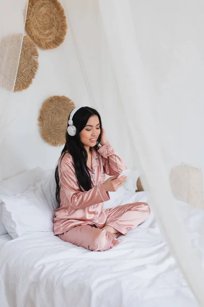 Positivo Joven Asiático Mujer Sentado Cama Con Auriculares Mirando Teléfono —  Fotos de Stock