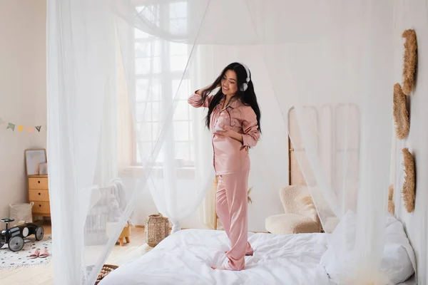Позитивная Молодая Азиатка Танцует Кровати Наушниках Держит Мобильный Телефон Спальне — стоковое фото