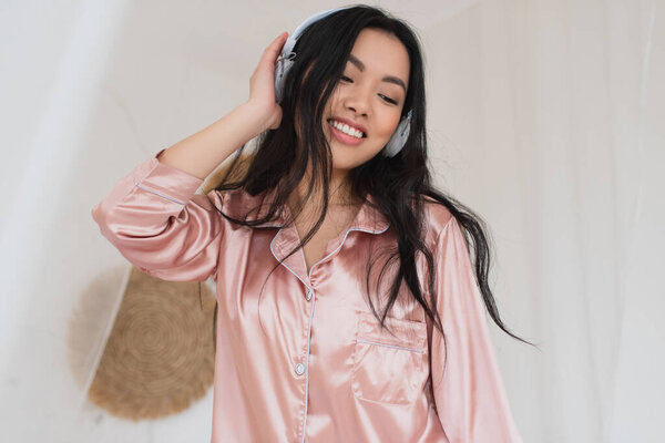 positive young asian woman in silk pajamas listening music with headphones in bedroom