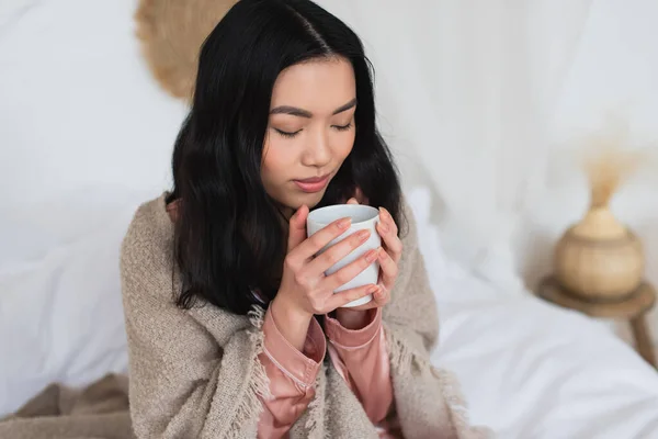 young asian woman in silk pajamas and blanket enjoying coffee aroma in bedroom