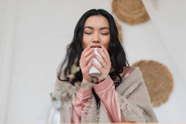 young asian woman holding cup of coffee in hands and enjoying aroma in bedroom