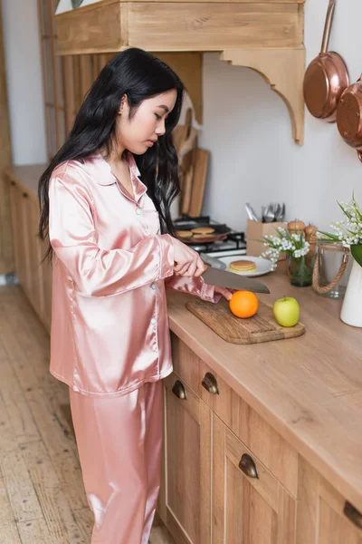 ピンクの絹のパジャマで台所で果物を切る若いアジアの女性 — ストック写真
