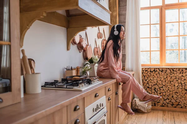 穿着粉色丝绸睡衣和耳机坐在炉边 在厨房里欣赏音乐的年轻亚洲女人微笑着 — 图库照片