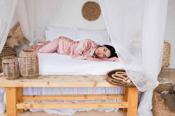 young asian woman in silk pajamas lying on white linen with outstretched hand in bedroom 