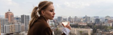 young woman holding glass of white wine and posing with cityscape on blurred background, banner clipart