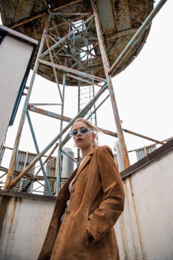 Mavi güneş gözlüklü modanın düşük açılı görüntüsü çatıdaki inşaata yakın cepte poz veriyor