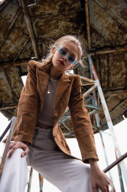 Mavi güneş gözlüklü güzel modelin düşük açılı görüntüsü ve çatıda inşaatın yanında poz veren modaya uygun kıyafet.