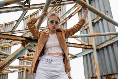 Mavi güneş gözlüklü genç model çatıda paslı yapının yanında poz veriyor.
