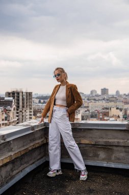 Mavi güneş gözlüklü modanın tam uzunluğu. Çatıda el ele poz vererek şehir manzarası ve bulanık ön plan.