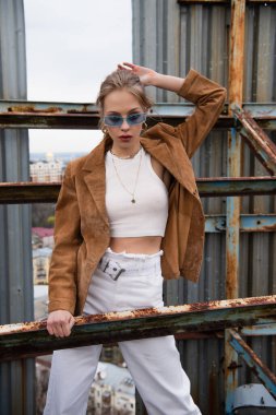 Sarışın, mavi güneş gözlüklü ve son moda elbiseli bir kadın çatıdaki yıpranmış yapının üzerinde duruyor. 