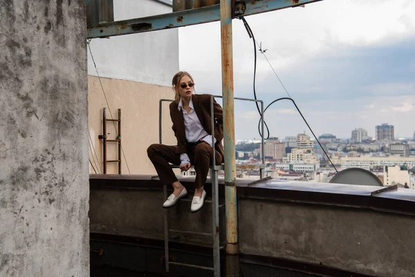 スタイリッシュなサングラスと屋上の金属製の階段の近くに座って流行のスーツの若いモデルの完全な長さ — ストック写真