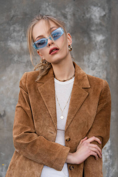 young woman in blue sunglasses and trendy outfit posing near grey wall