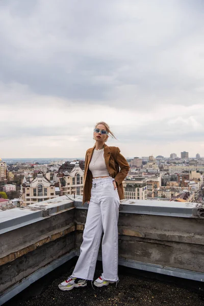 Tam boy mavi güneş gözlüklü genç model cepleri kapalı poz şehir manzaralı ve bulanık ön plan