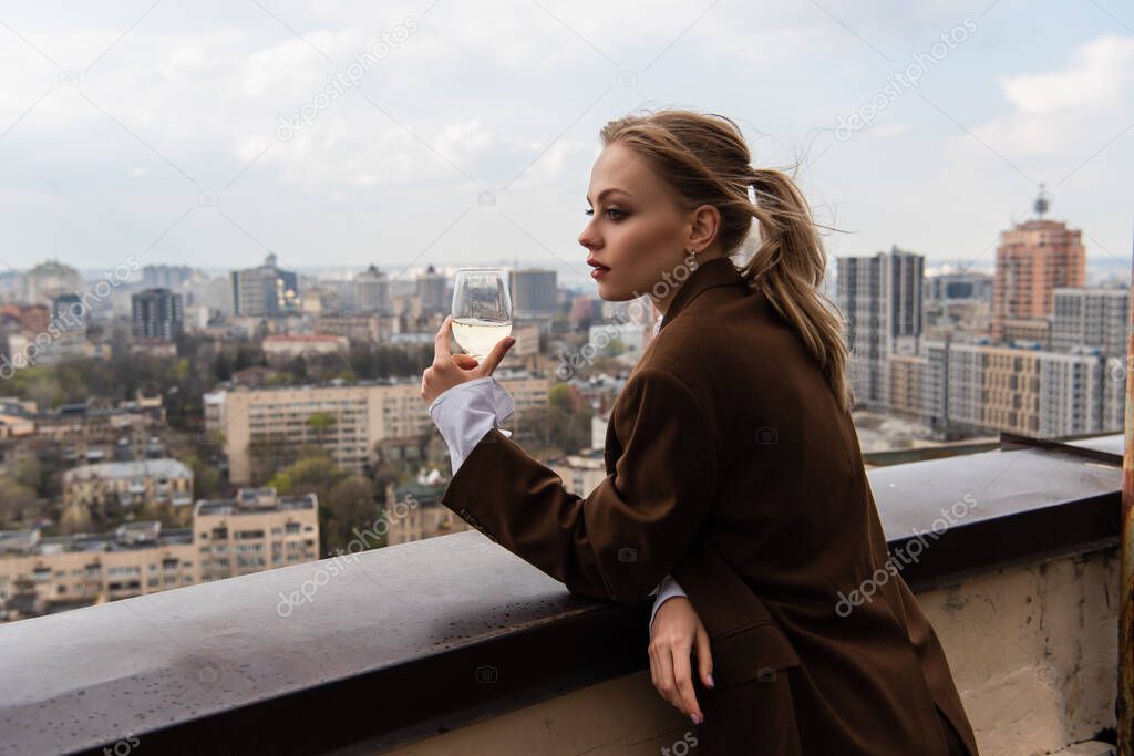 young woman holding glass of white wine and posing with cityscape on blurred background