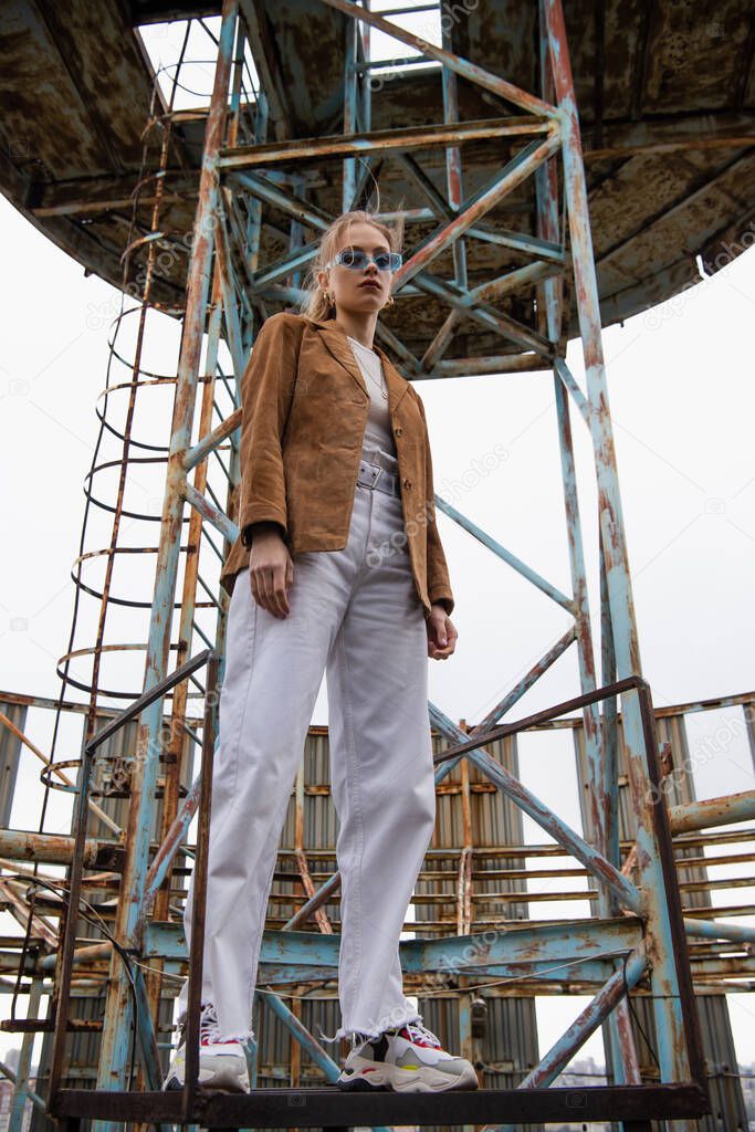 low angle view of pretty young model in blue sunglasses, white pants and suede jacket posing near construction on rooftop