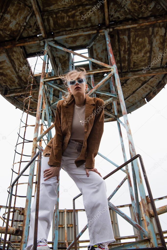 low angle view of model in blue sunglasses, white pants and suede jacket posing near construction on rooftop