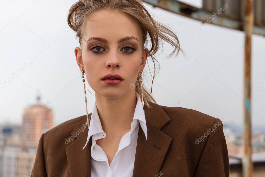 young model in earrings, white shirt and brown jacket looking at camera on rooftop 