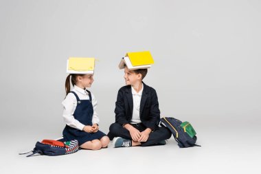 happy schoolkids in uniform with books on heads sitting and looking at each other on grey clipart