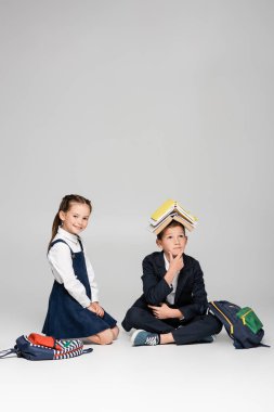 pensive schoolboy with books on head sitting near cheerful girl on grey clipart