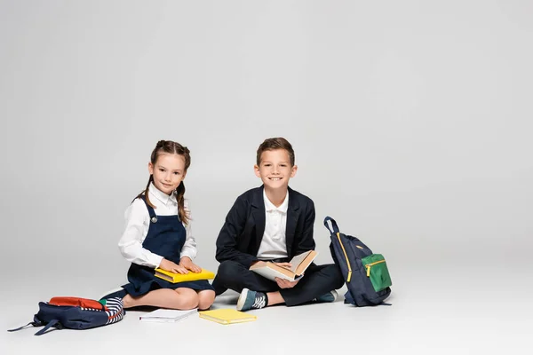 有名な制服を着た幸せな子供たちがバックパックや本とグレーで座っています — ストック写真