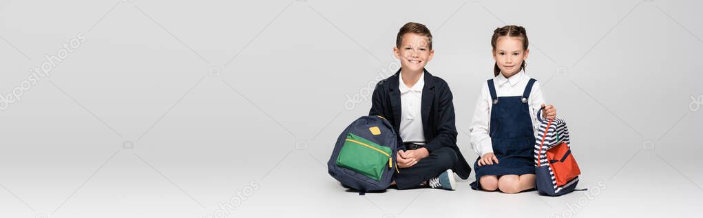 happy schoolkids in prestigious uniform sitting with backpacks on grey, banner