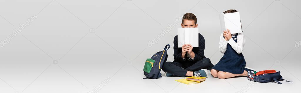 schoolkids in uniform sitting and covering faces with books near backpacks on grey, banner