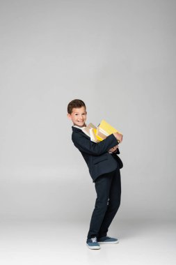 happy schoolboy in blazer and pants holding pile of books on grey clipart