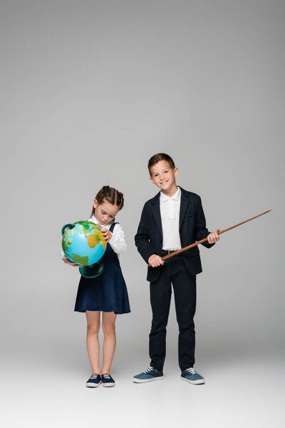 веселый школьник, держащий палку рядом с грустной девушкой в платье с глобусом на сером 