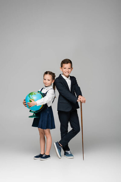 smiling schoolboy holding pointing stick near girl in dress with globe on grey 