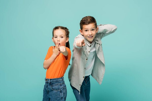 shocked girl in orange t-shirt covering mouth near boy pointing at camera isolated on blue