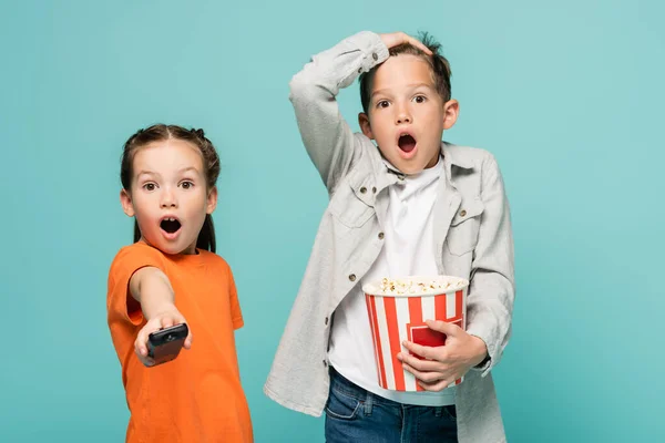 Geschokt Meisje Houden Afstandsbediening Buurt Jongen Met Popcorn Emmer Geïsoleerd — Stockfoto