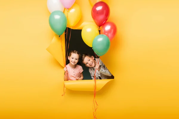 Veselé Děti Drží Barevné Balónky Přes Díru Žlutém Pozadí — Stock fotografie