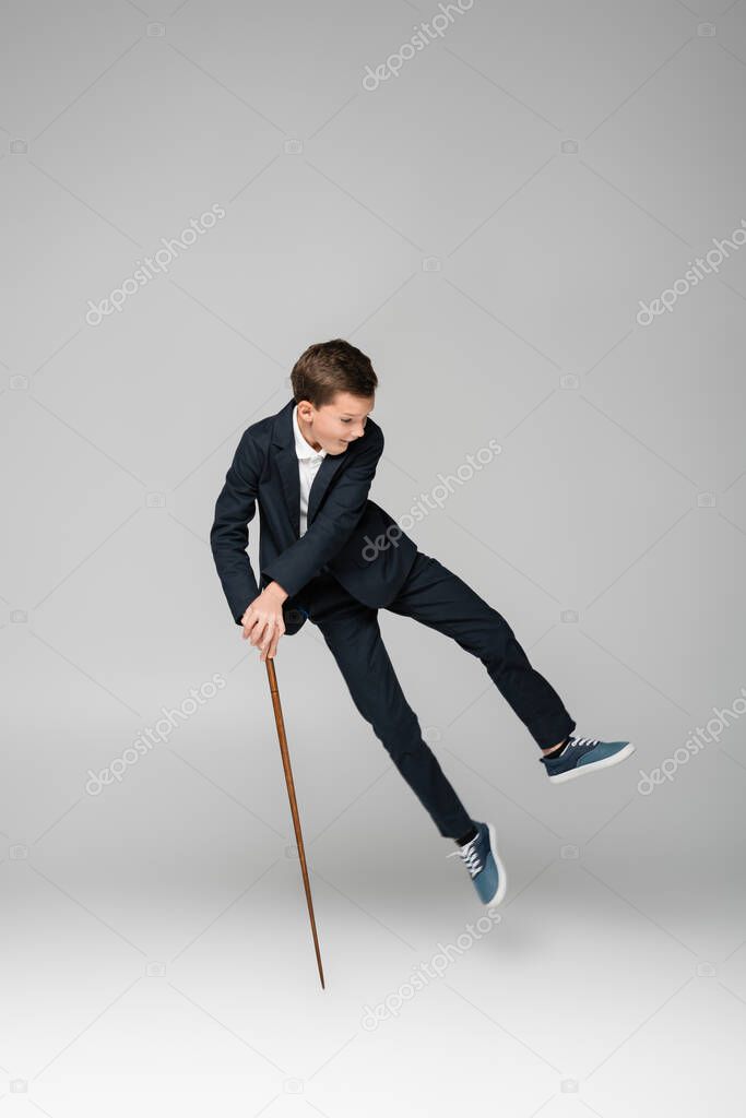 amazed schoolboy in pants and blazer jumping with pointing stick on grey